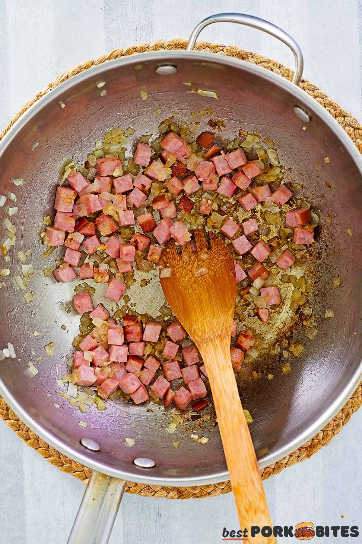 a pan with sauted onions, garlic and ham, being stirred with a wooden spoon