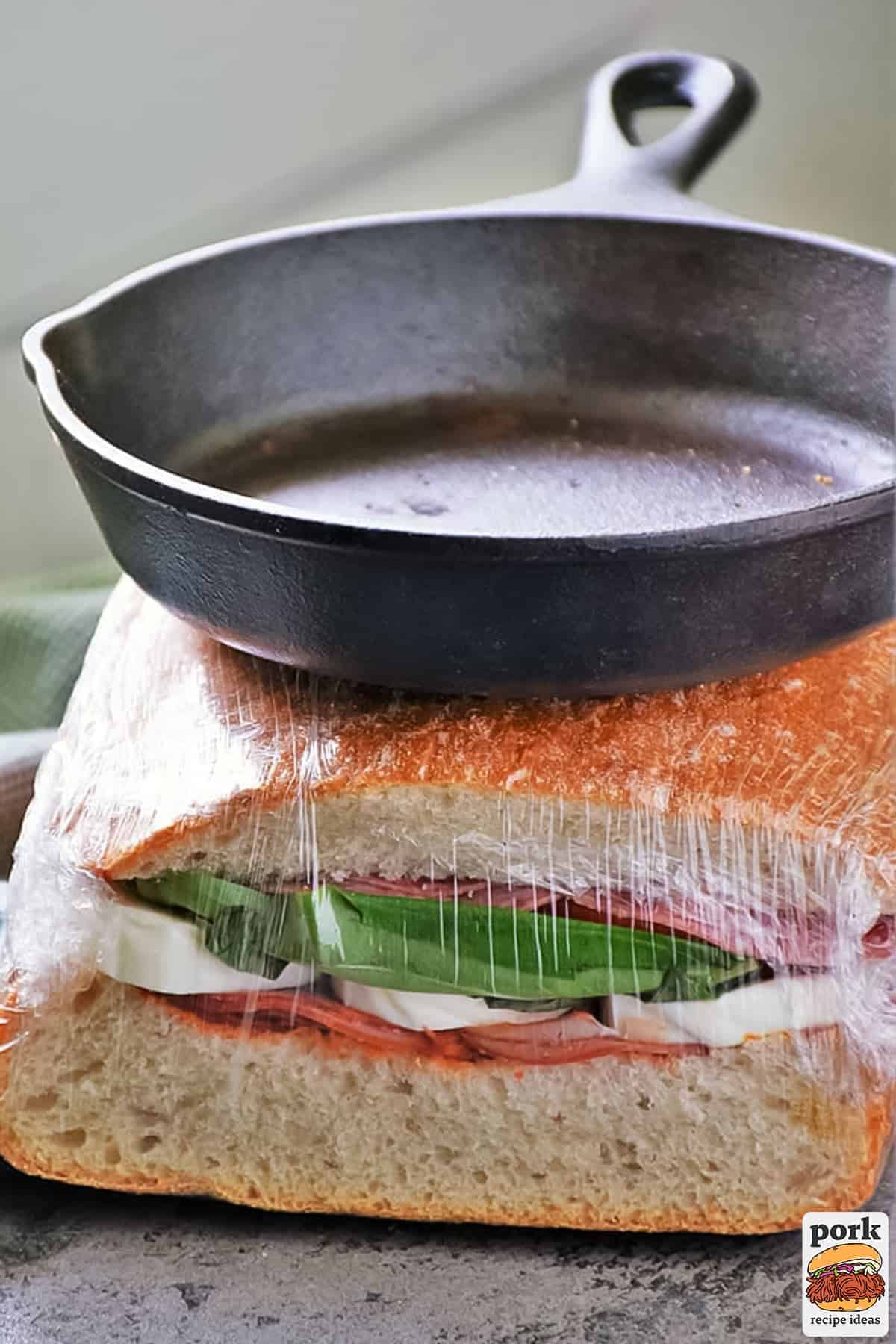 an italian sandwich wrapped in plastic and being pressed under a cast iron pan