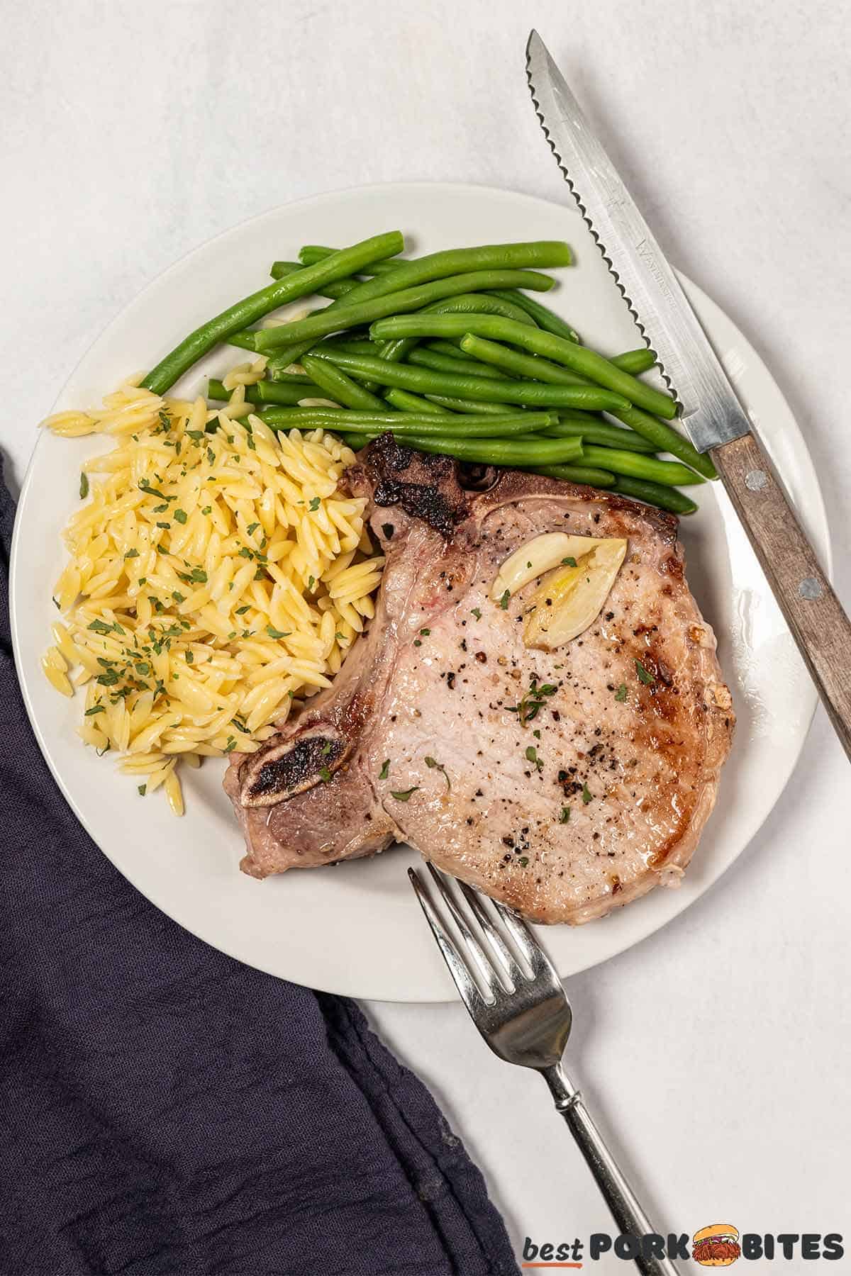 pan fried pork chop on a white plate next to orzo and green beans with a fork and knife