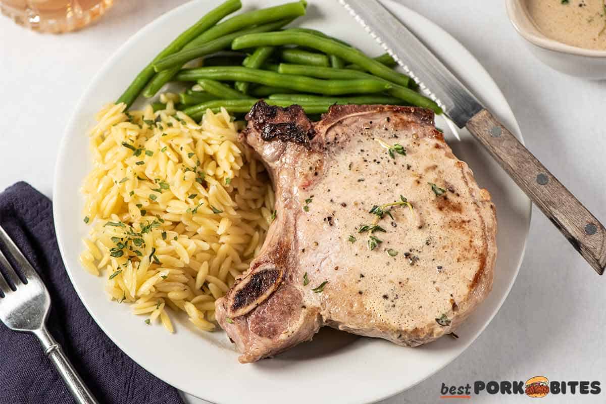 pan fried pork chop up close on a white plate with a knife, orzo, and green beans
