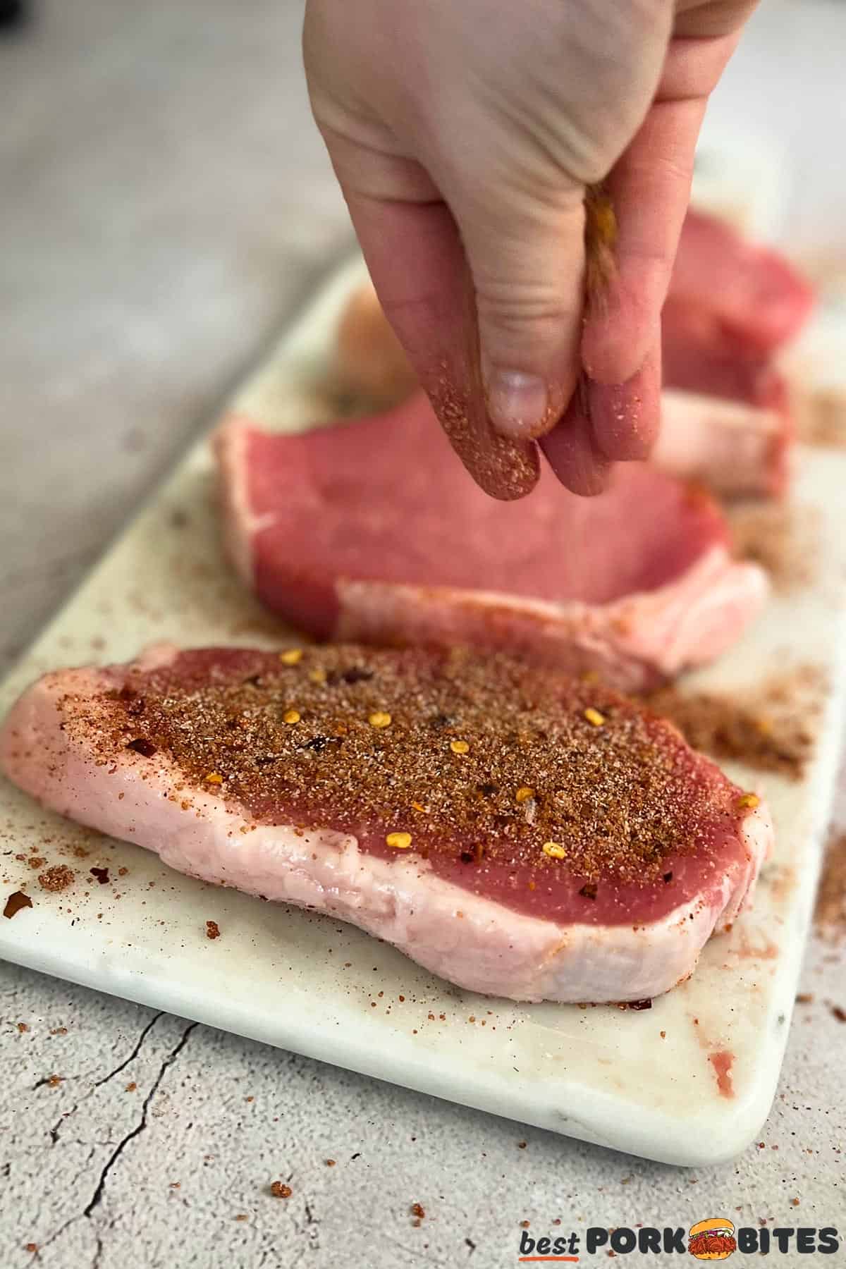raw pork chops having pork chop rub sprinkled onto them on a white cutting board