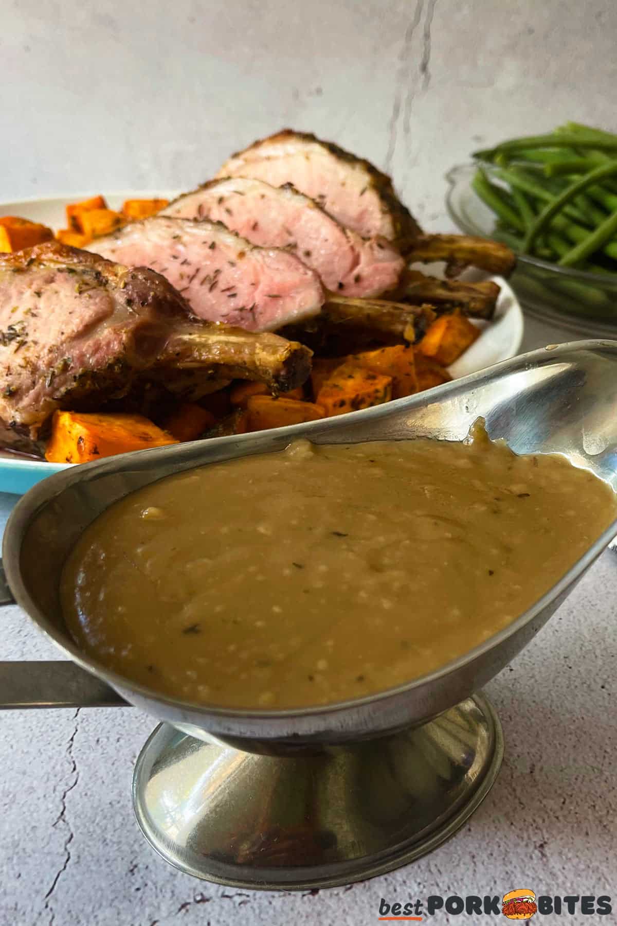 a saucer of gravy in front of a dish of pork chops, sweet potatoes and green beans
