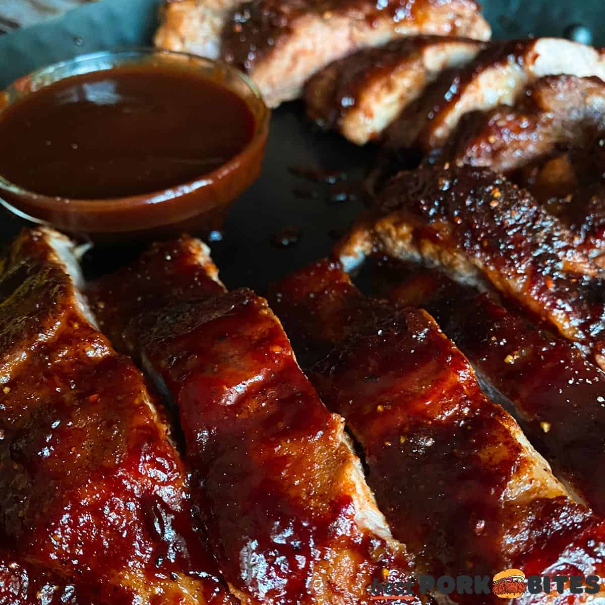 a closeup of sliced and cooked pork ribs on a black plate with a bowl of sauce