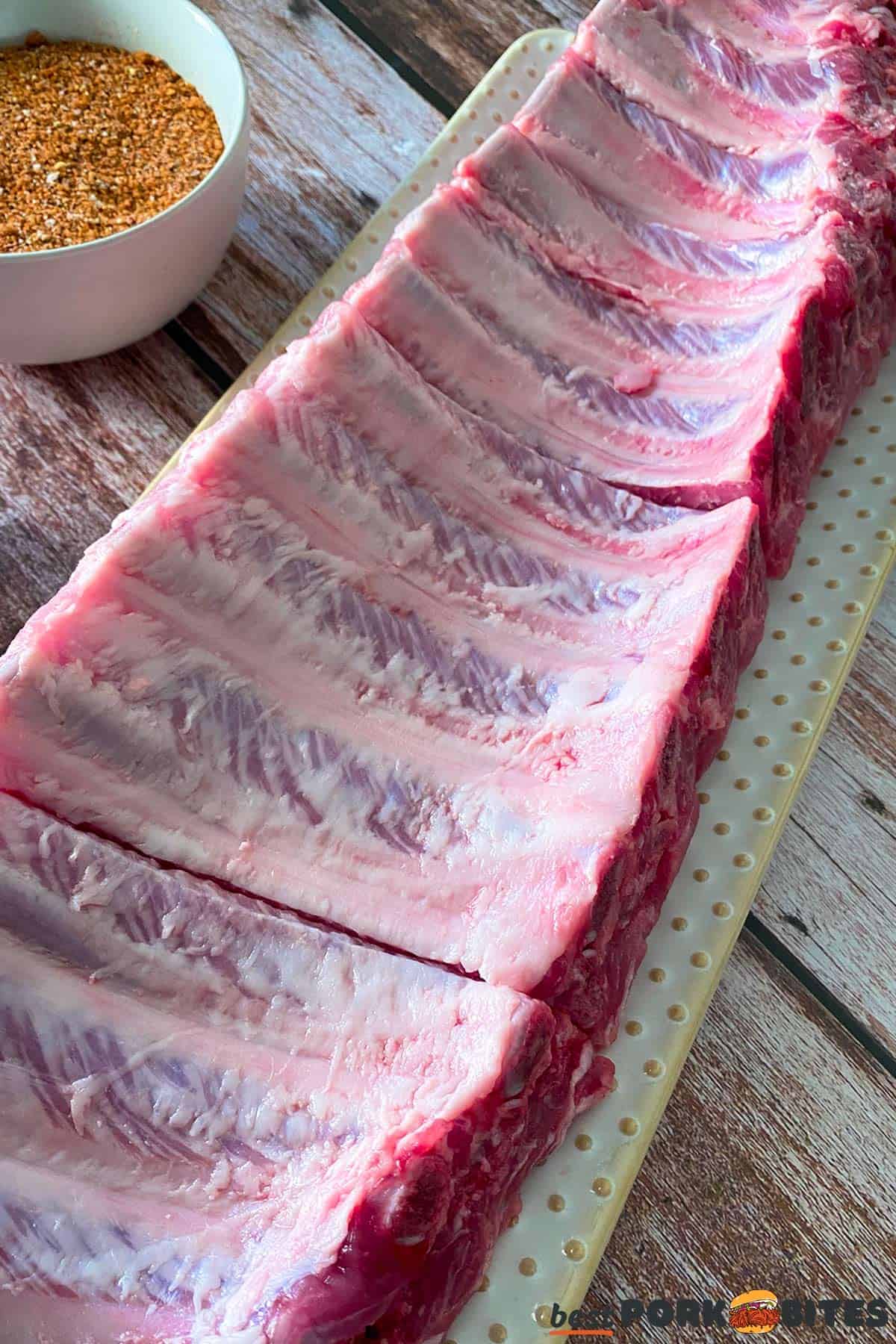 raw pork ribs on a cutting board, cut into four equal parts, next to a bowl of rib rub