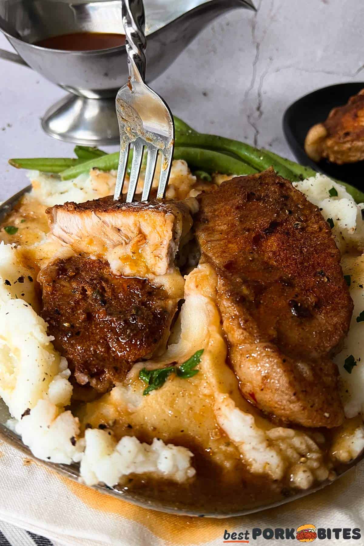 a cut open pork chop being held up by a fork on a plate of mashed potatoes, green beans and gravy