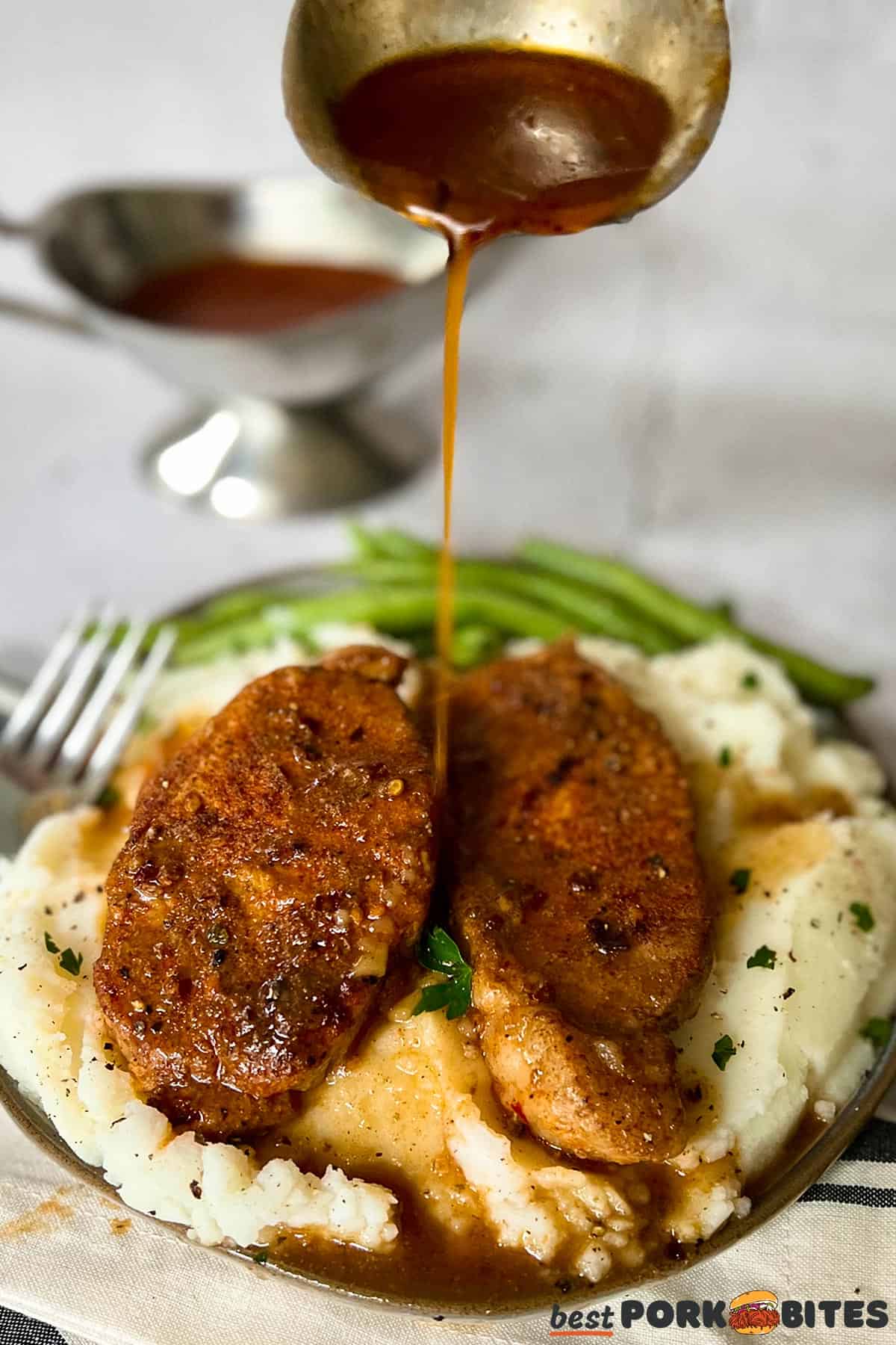gravy being poured from a spoon on to a plate of mashed potatoes, pork chops and green beans