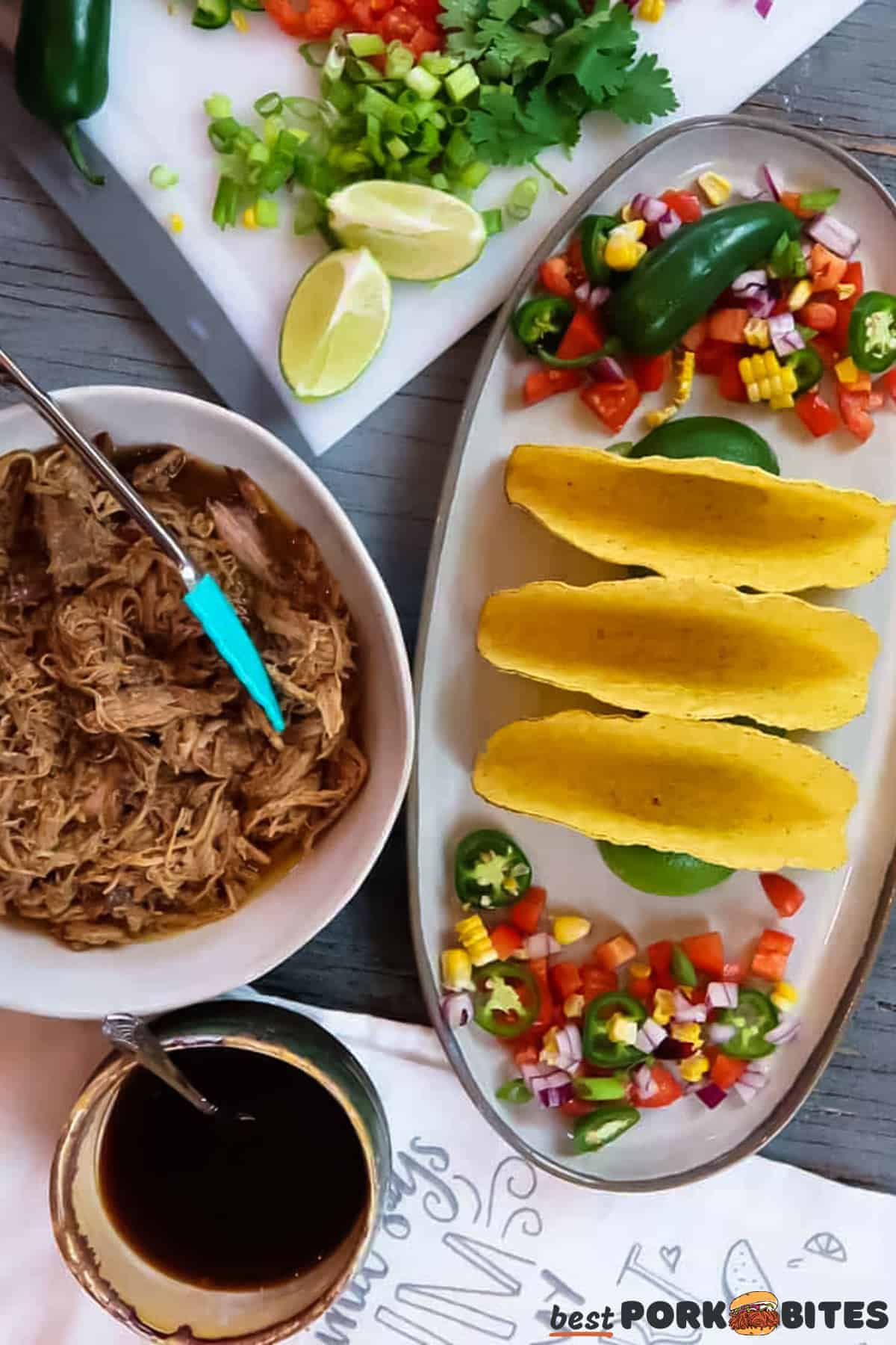 a plate of taco shells with sliced toppings next to a cutting board of toppings, a bowl of pulled pork and a dish of sauce