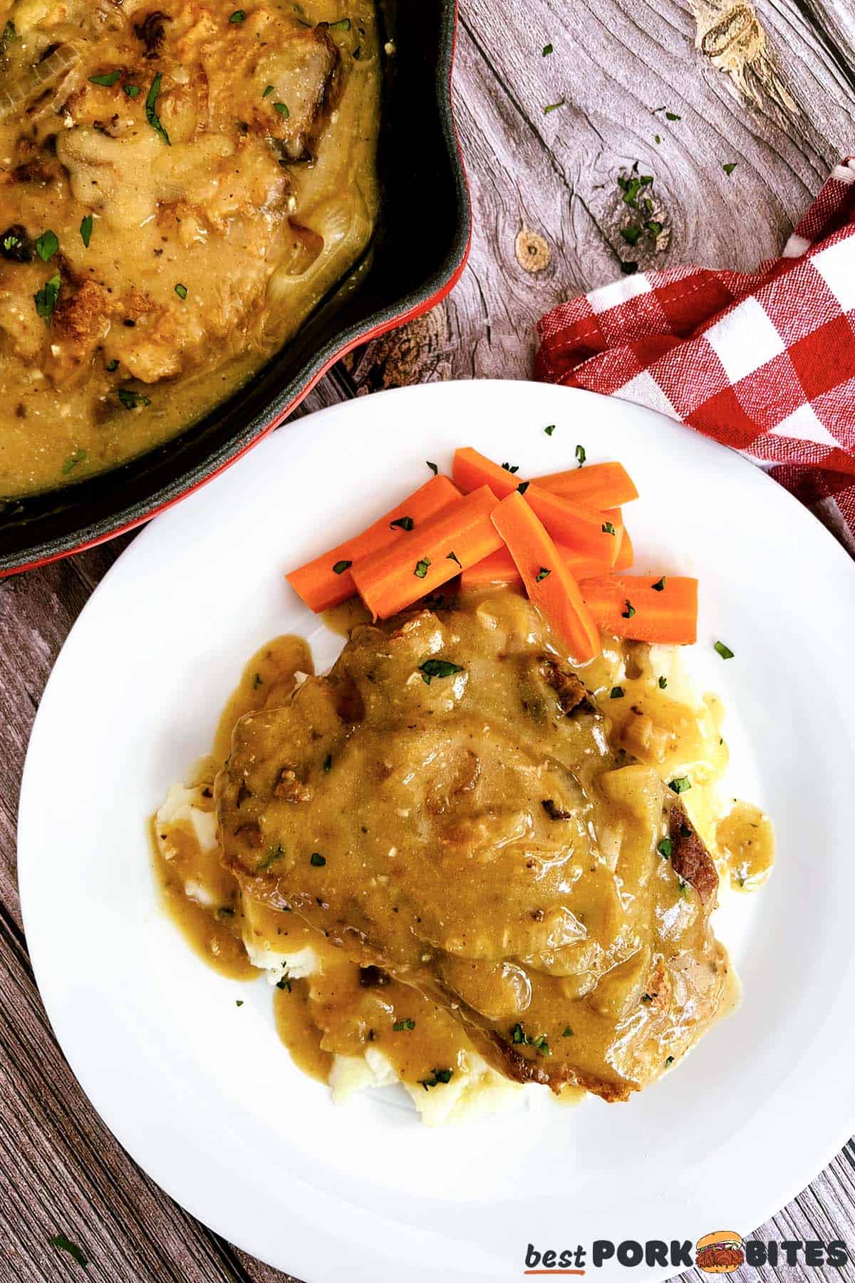 smothered pork chops on a plate with carrots and mashed potatoes next to the skillet of gravy